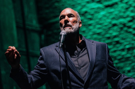 Roger Guenveur Smith, in a pinstriped suit, raises one hand at the microphone against a green-lit brick wall