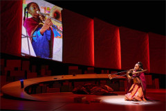 A girl kneels with a trombone beneath a projected image, in red light in creation stories and all the important importants. Photo by Valerie Terranova