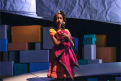 A girl clutches a red blanket among stacks of colored boxes in creation stories and all the important importants. Photo by Valerie Terranova