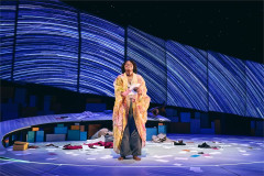 A woman in a patterned robe stands alone at center stage beneath streaks of light in creation stories and all the important importants. Photo by Valerie Terranova