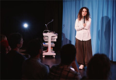 Rachel Kaly addresses the audience in front of a blue curtain in Hospital Hour. Photo by Emilio Madrid
