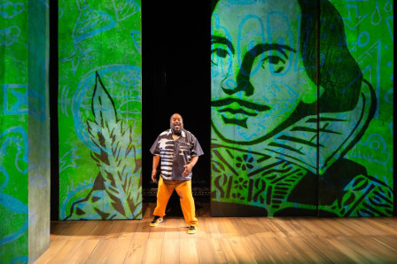 Jacob Ming-Trent alone on a wooden stage in front of tall panels projected with a green portrait of Shakespeare