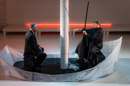 Two men in black sit in a white paper rowboat beneath a pillar rigged as a mast, one holding a pole, in The Invention of Love at Theaterlab