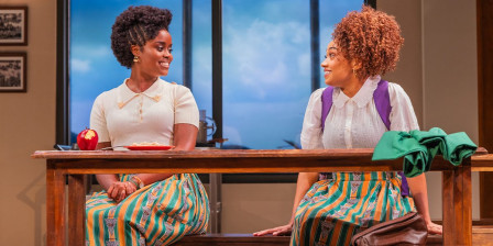 Denee Benton and Jasmine Amy Rogers sit facing each other across a wooden cafeteria table in green and orange school uniforms