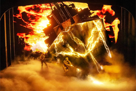 A vast structure breaks apart in fire and forked lightning above a fogbound stage, two figures below it. Photo by Matthew Murphy and Evan Zimmerman