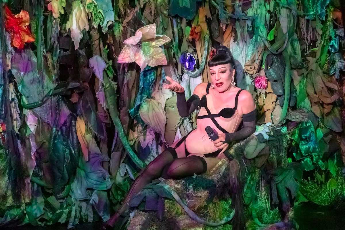 Joey Arias reclining in a dense fabric jungle of oversized flowers and vines, microphone in hand