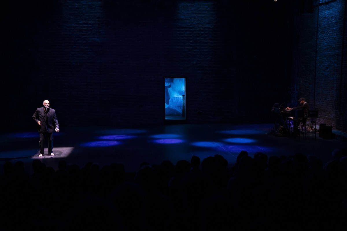 Wide view from the auditorium: Smith in a pool of blue light at the left of the bare stage, musician Marc Anthony Thompson at the right