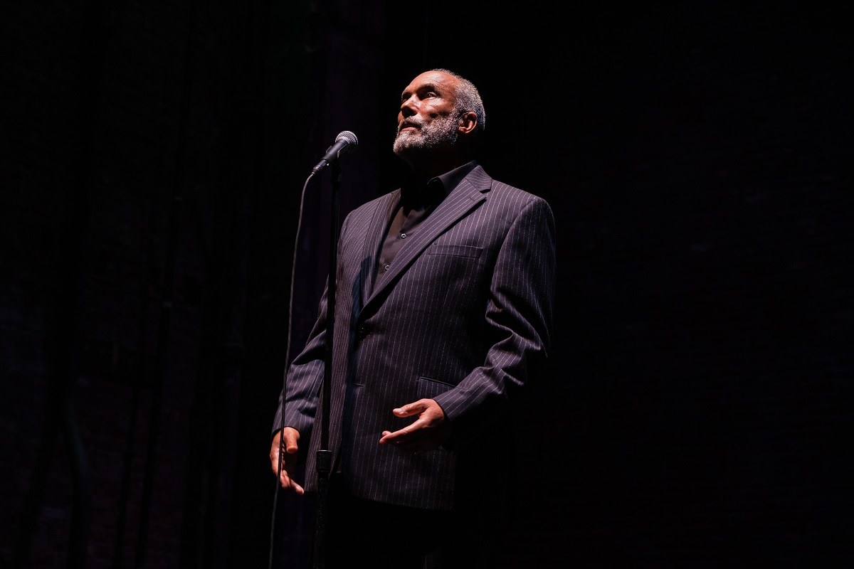 Smith at the microphone in a tight spotlight, surrounded by darkness, his head turned upward