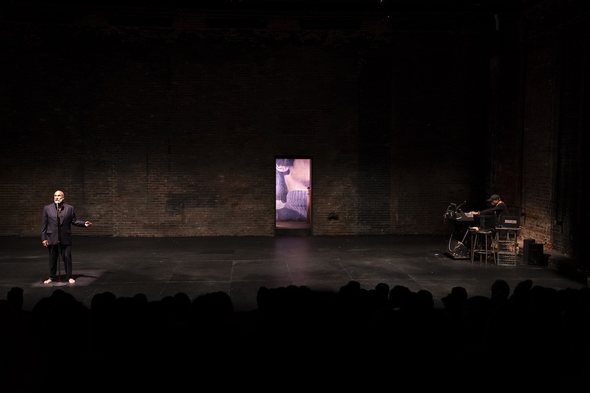 The stage seen over the heads of the audience, a lit panel at the centre, Smith at the left and his musician at the right