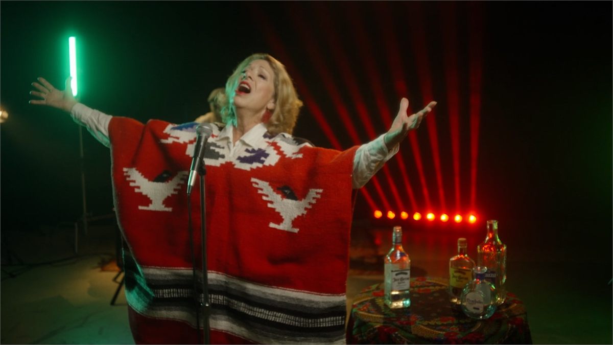 Stephanie Trudeau, arms spread wide in a red and black jorongo at a standing microphone, lit by green neon and a red backlight, with tequila bottles on a table beside her