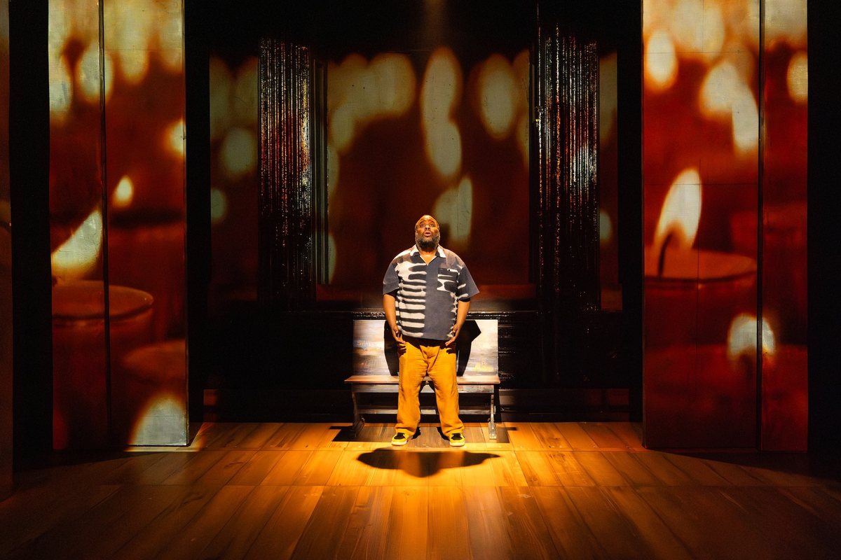 Jacob Ming-Trent seated alone on a wooden bench, lit warmly against amber projections