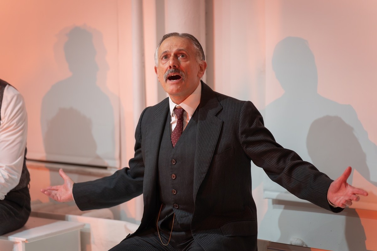 David Greenspan as A. E. Housman, arms spread in a three-piece suit against the white wall of Theaterlab