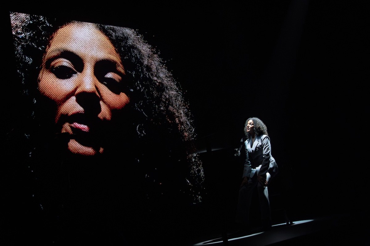 Sarah Jones performs beside her own face in close-up on the back projection screen in America, Who Hurt You?. Photo by HanJie Chow Sarah Jones performs beside her own face in close-up on the back projection screen in America, Who Hurt You?. Photo by HanJie Chow