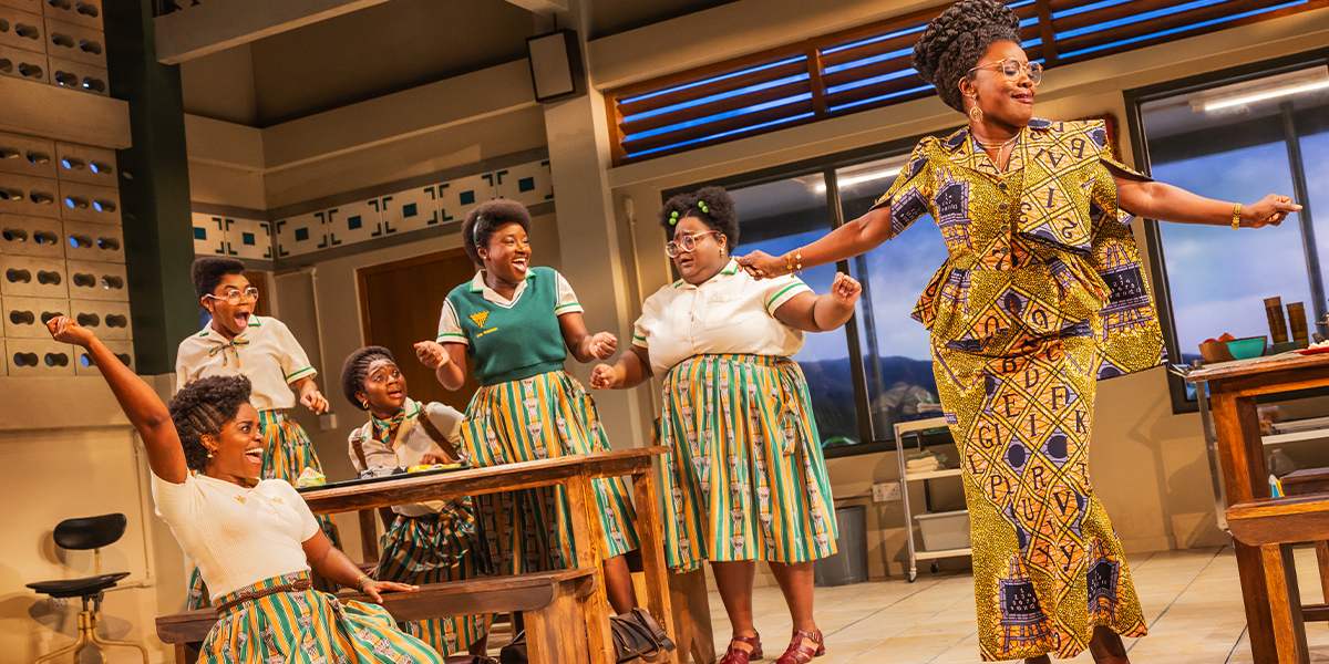 The company of School Girls cheer around a cafeteria table as the headmistress dances in an alphabet-print dress