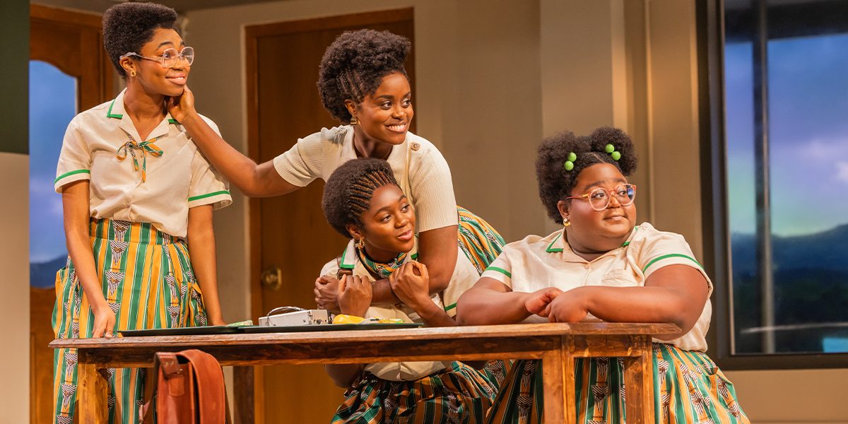 Four students in green and orange school uniforms lean together over a desk, laughing